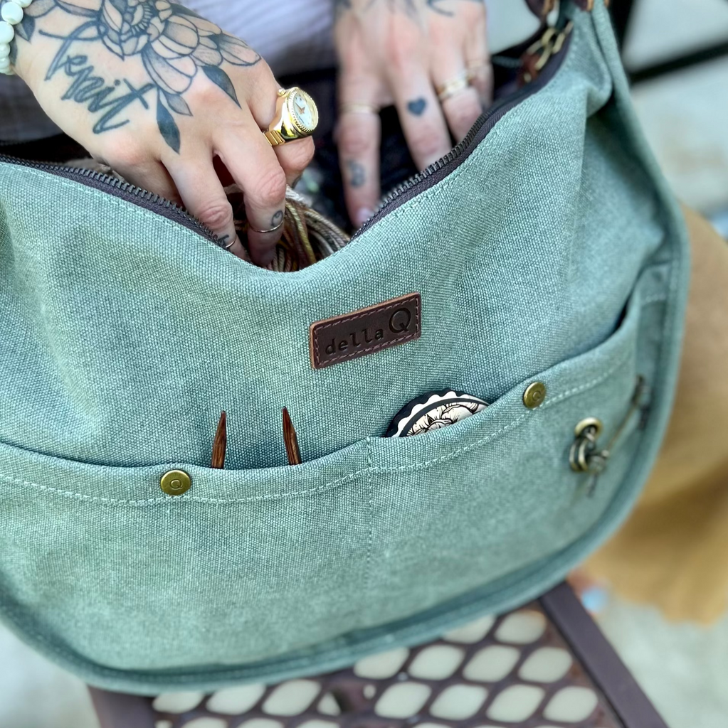 Close-up of a light green adella Q Everyday Maker’s Bag being opened by a person with floral and script tattoos on their hands. Knitting needles and accessories peek out of the front pocket.